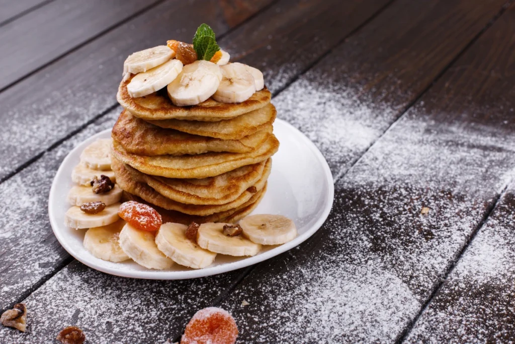 A stack of fluffy buckwheat banana pancakes drizzled with maple syrup, topped with banana slices and chopped nuts, served on a rustic wooden plate with a cozy breakfast background."