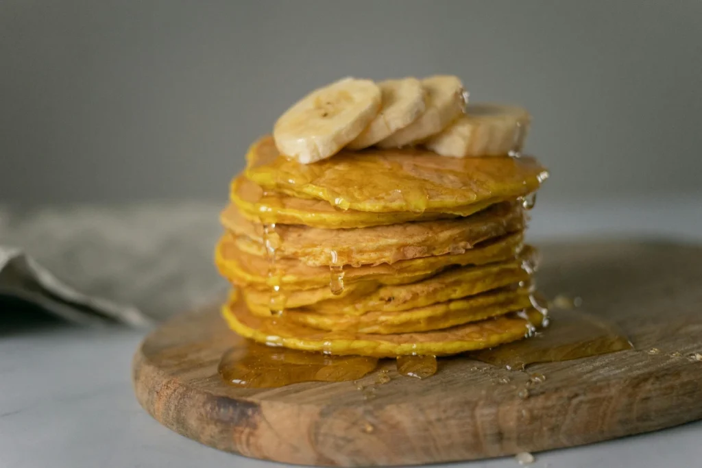 A stack of fluffy buckwheat banana pancakes drizzled with maple syrup, garnished with banana slices and nuts, served on a rustic wooden plate with a cozy breakfast background.