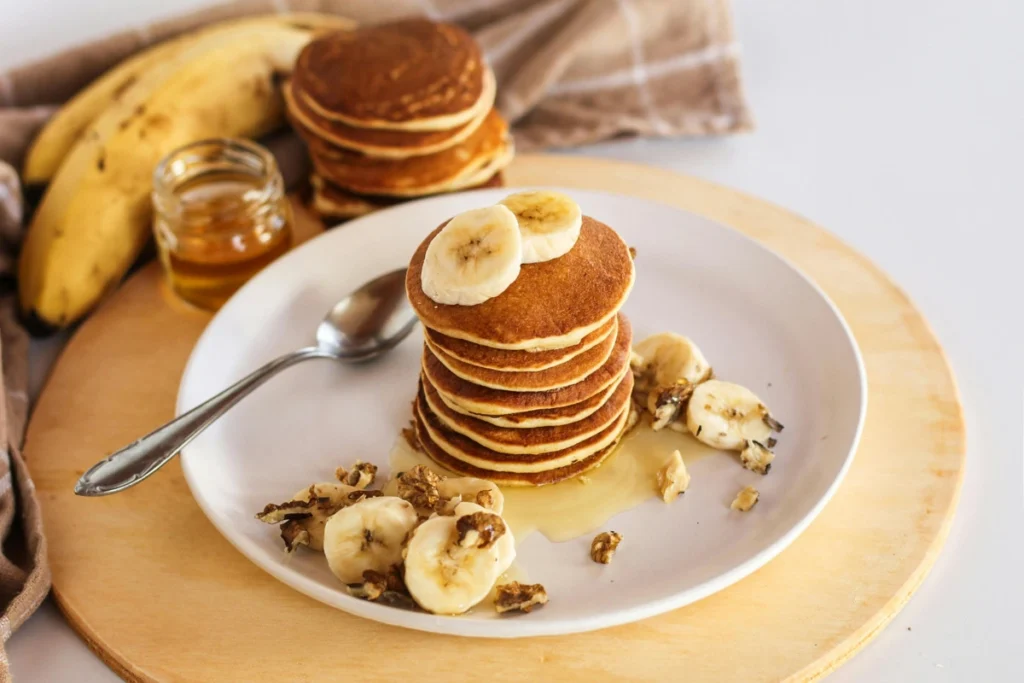 A close-up of golden-brown buckwheat banana pancakes with syrup cascading down the sides, served with fresh bananas and a sprinkle of chopped nuts on a wooden table.