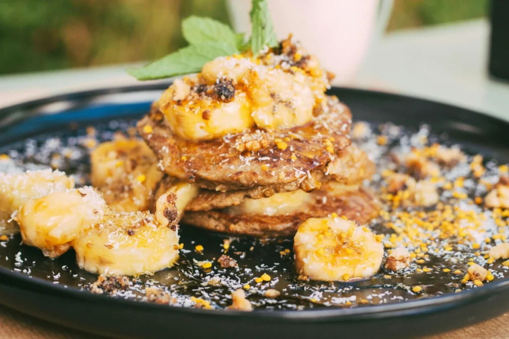 A stack of fluffy buckwheat banana pancakes drizzled with maple syrup, garnished with banana slices and nuts, served on a rustic wooden plate with a cozy breakfast background."